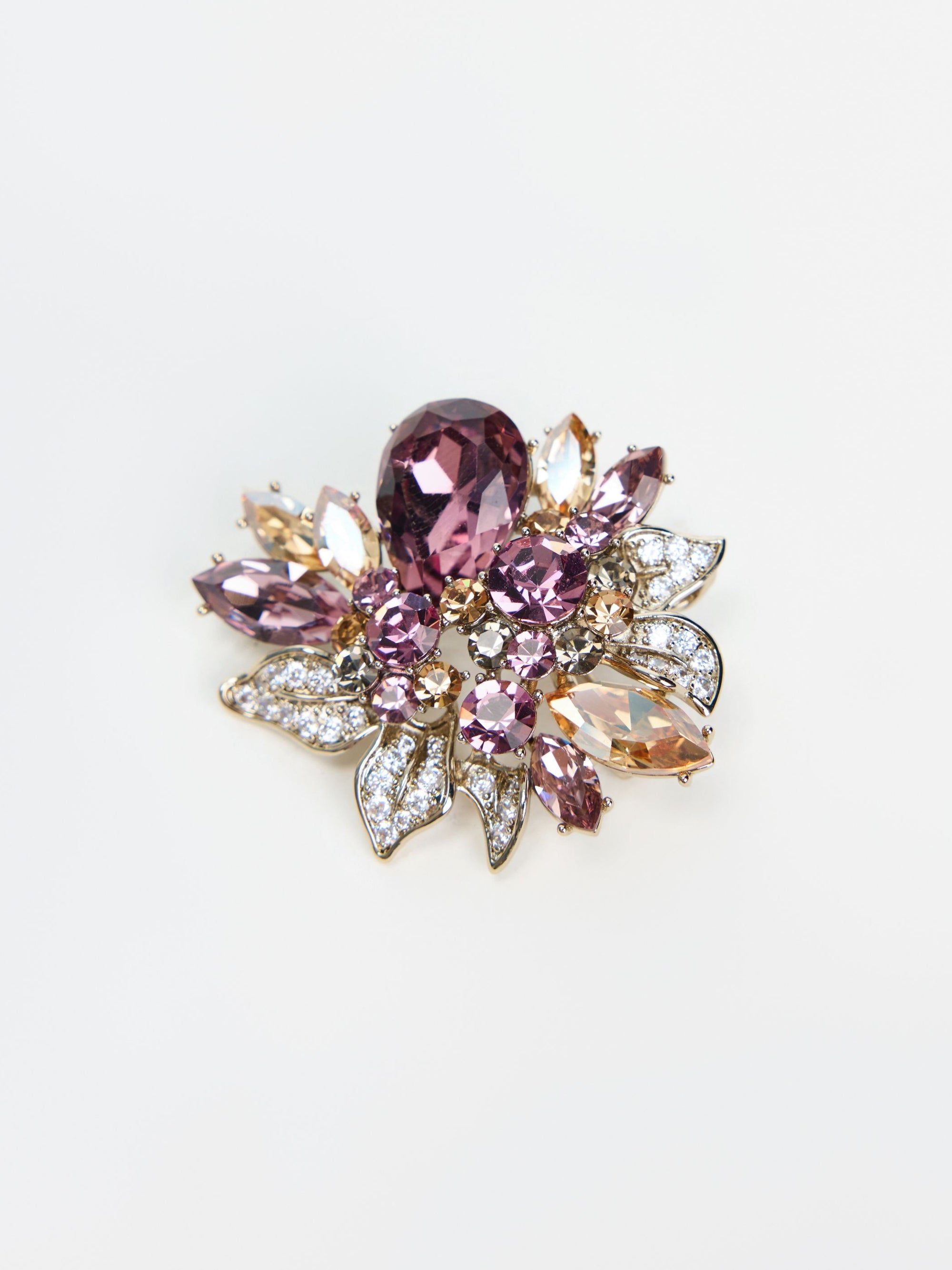 The Social Queen Amethyst Floral Brooch showcases amethyst-toned crystals and clear gemstones arranged in an elegant floral motif, highlighted by a gold-tone finish with silver accents, all set against a white background.