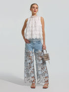 A woman stands against a plain background wearing the Social Queen 3D Floral Lace Appliqué Silk Top paired with blue jeans featuring a sheer lace overlay, styled with a small purse and open-toed heels.