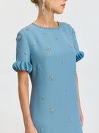 A woman wears the Social Queen Beaded Crystal Embellished Dress, featuring a light blue shift silhouette with jeweled floral details and pleated ruffle cuffs, standing against a plain background.