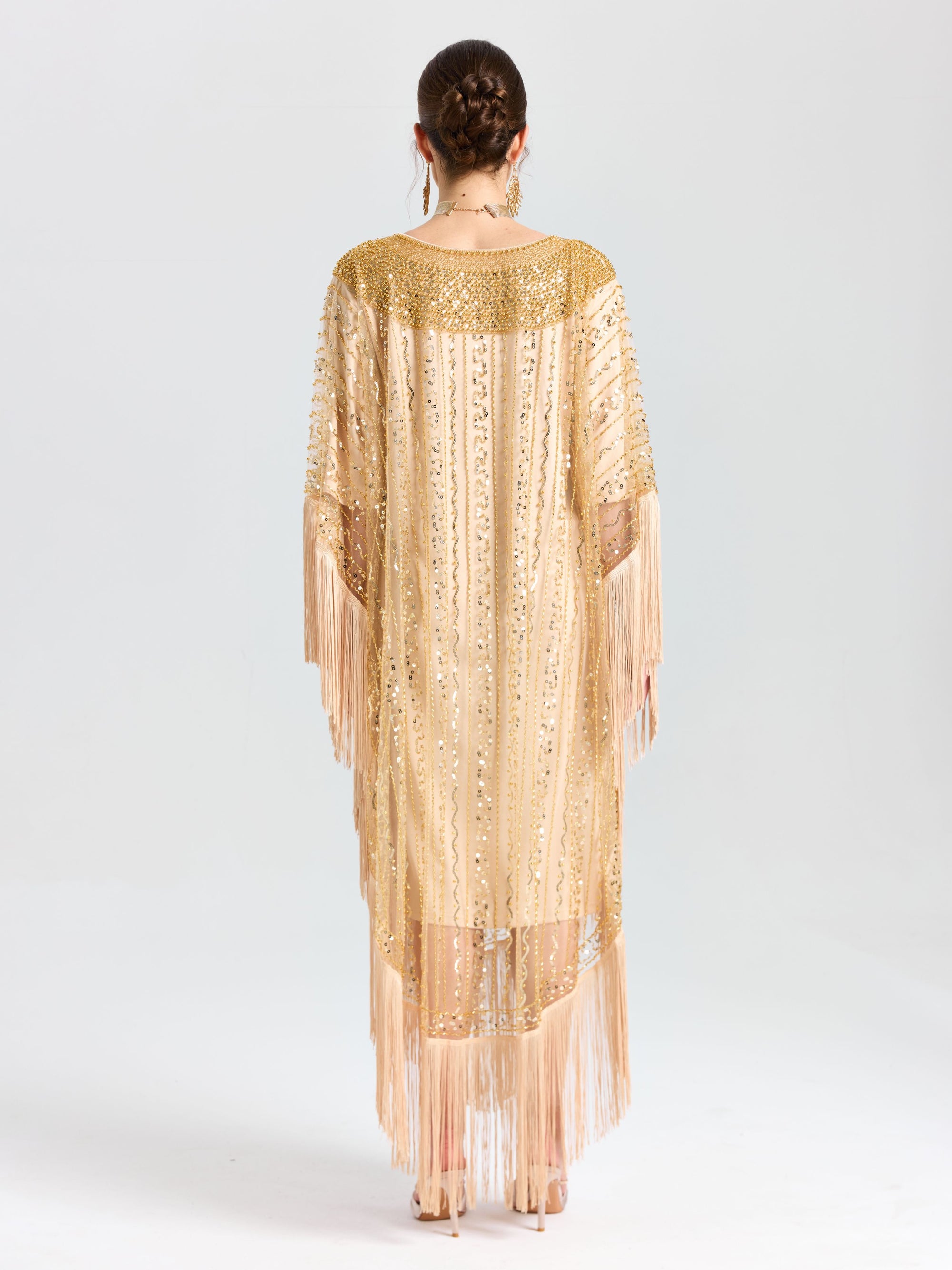 A woman faces away, wearing the Social Queen Beaded Fringe Cape Dress featuring gold beaded sequins, fringe trim, and sheer panels. The background is plain white.