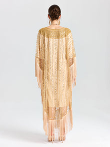 A woman faces away, wearing the Social Queen Beaded Fringe Cape Dress featuring gold beaded sequins, fringe trim, and sheer panels. The background is plain white.