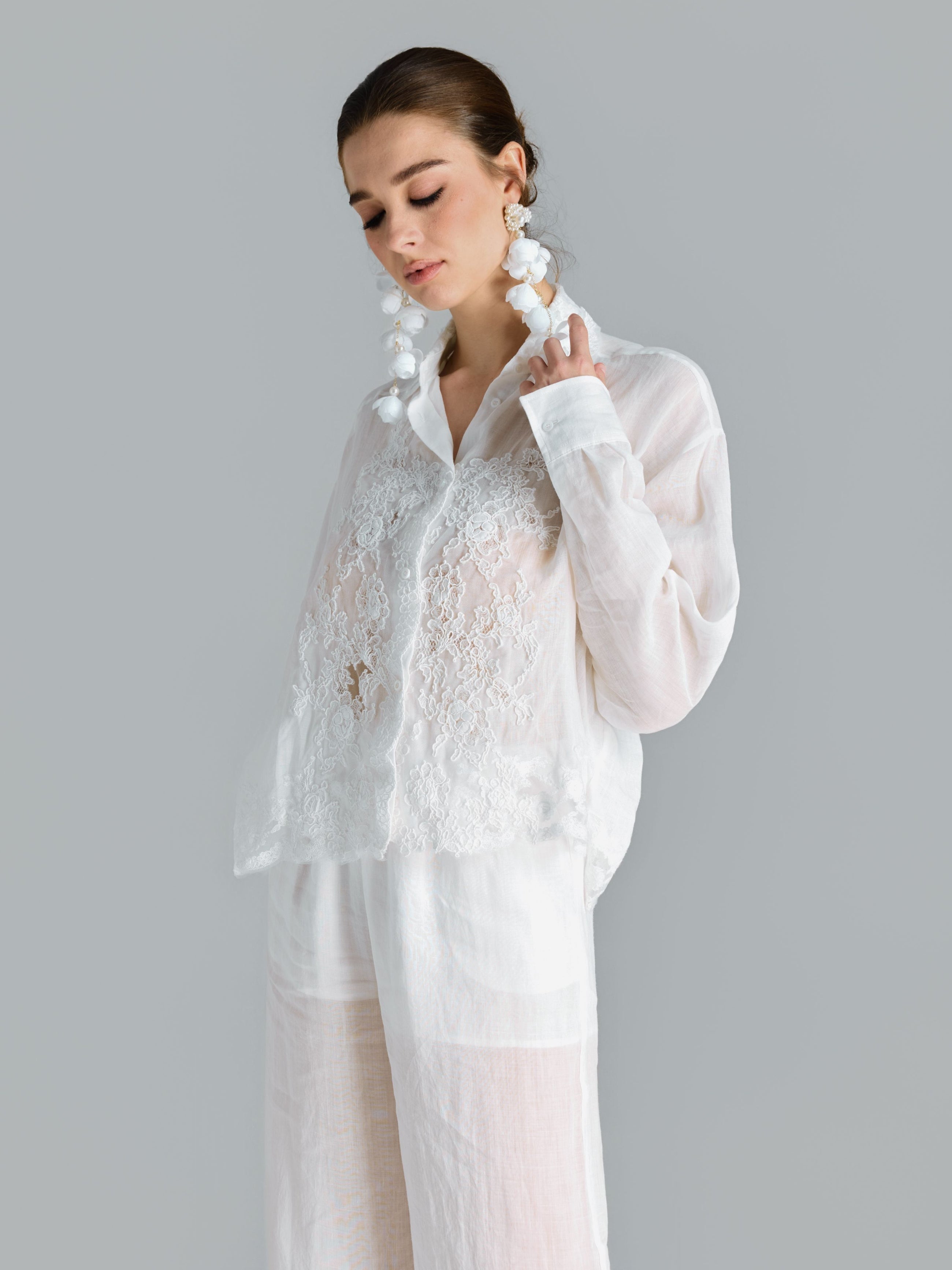 A woman stands against a plain gray background, looking down with her hand near her face, wearing the Social Queen 3D Lace Appliqué Shirt with a relaxed silhouette, paired with white pants and large white earrings.