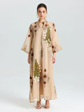 A woman faces forward in the Social Queen Placement Embroidered Linen Dress, a long beige piece featuring floral and leaf embroidery with sequin details, paired with open-toed heels against a plain light background.