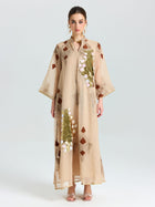 A woman faces forward in the Social Queen Placement Embroidered Linen Dress, a long beige piece featuring floral and leaf embroidery with sequin details, paired with open-toed heels against a plain light background.