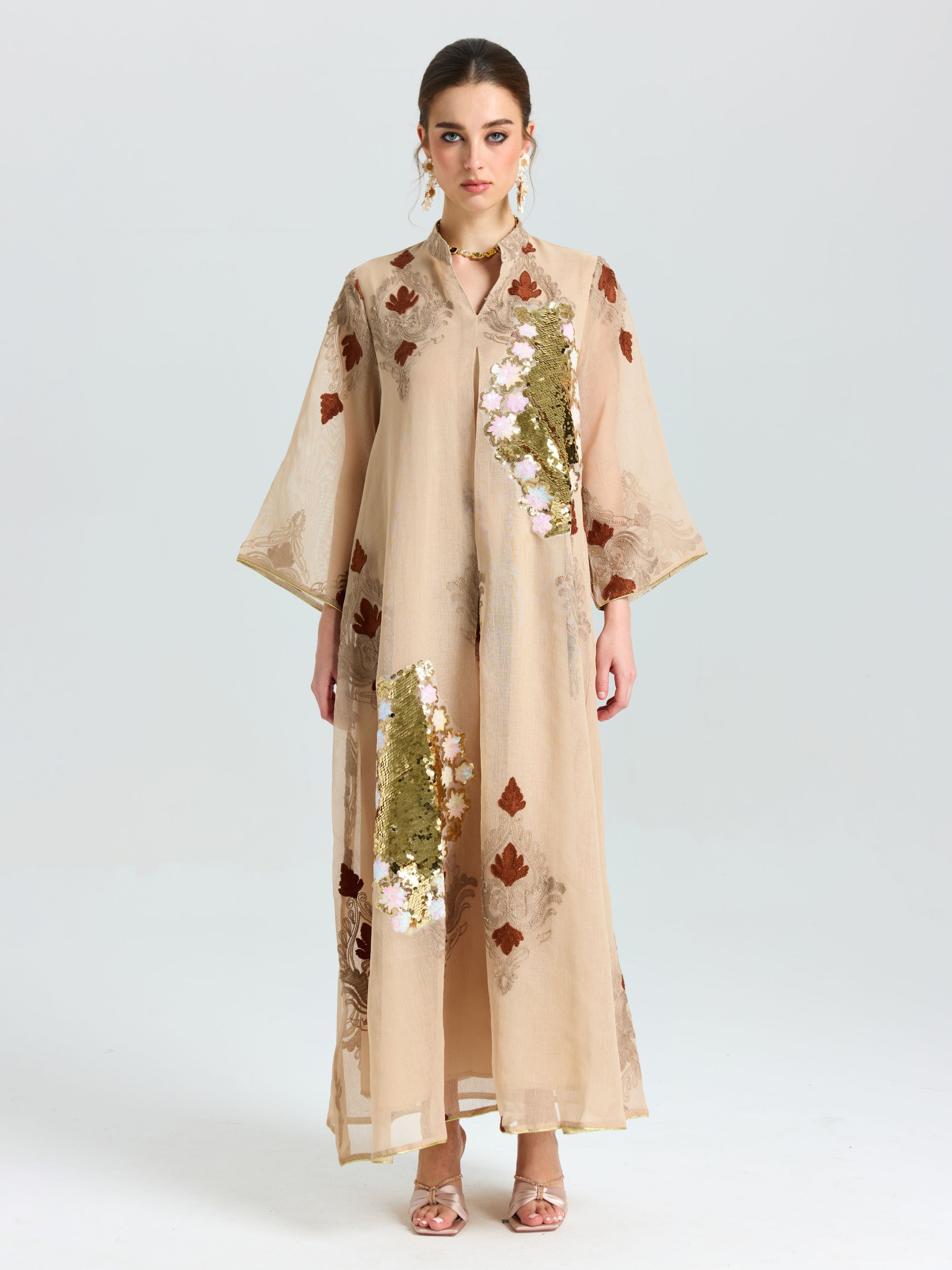 A woman faces forward in the Social Queen Placement Embroidered Linen Dress, a long beige piece featuring floral and leaf embroidery with sequin details, paired with open-toed heels against a plain light background.