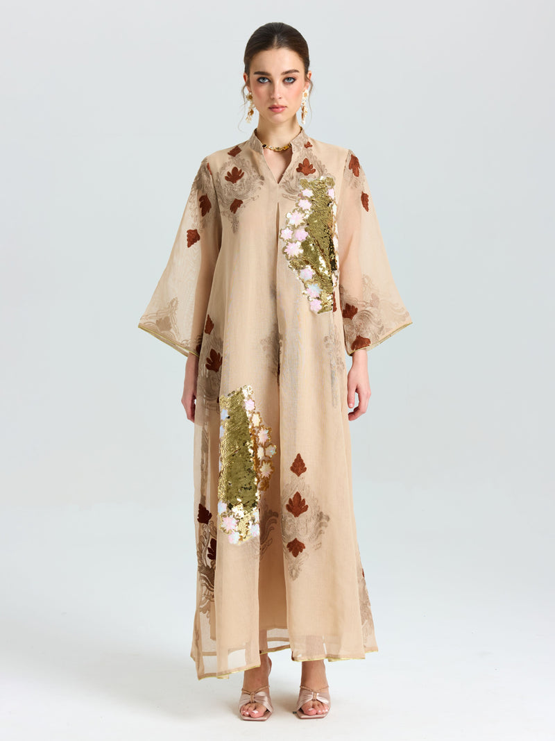 A woman faces forward in the Social Queen Placement Embroidered Linen Dress, a long beige piece featuring floral and leaf embroidery with sequin details, paired with open-toed heels against a plain light background.