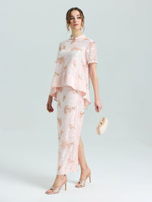 A woman wears the Dragonfly Beaded Lace Top by Social Queen—a light pink, asymmetrical lace top with beaded embroidery and short sleeves—paired with a small purse and high-heeled sandals, perfect for special occasions.