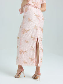 A person models the Social Queen Dragonfly Beaded Lace Midi Skirt—light pink with a floral design, straight silhouette, and back slit—paired with nude heeled sandals against a plain background.