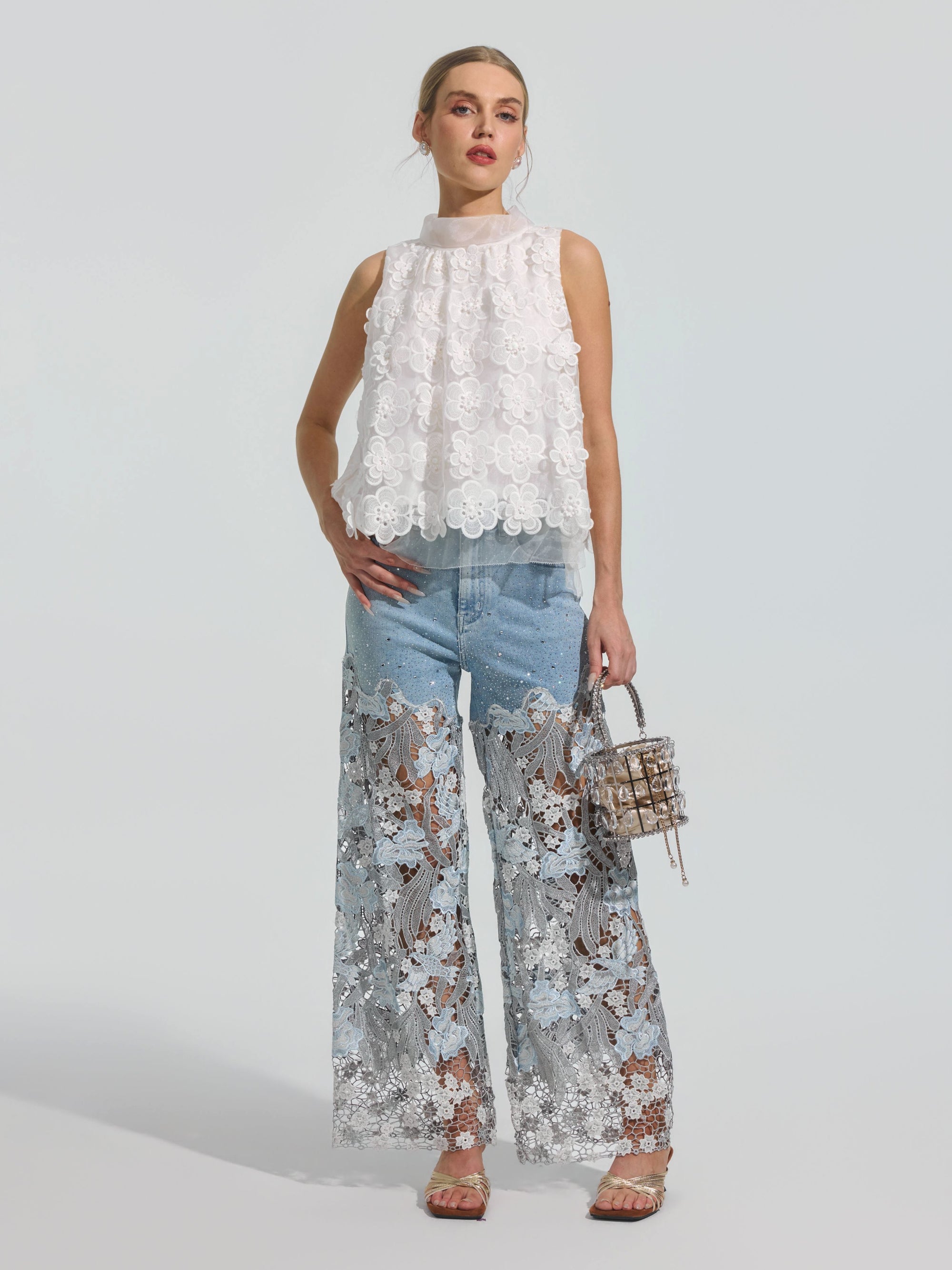 A woman stands against a plain background wearing Social Queen’s Floral Lace Patchwork Pants, styled with a sleeveless white top and a small handbag.
