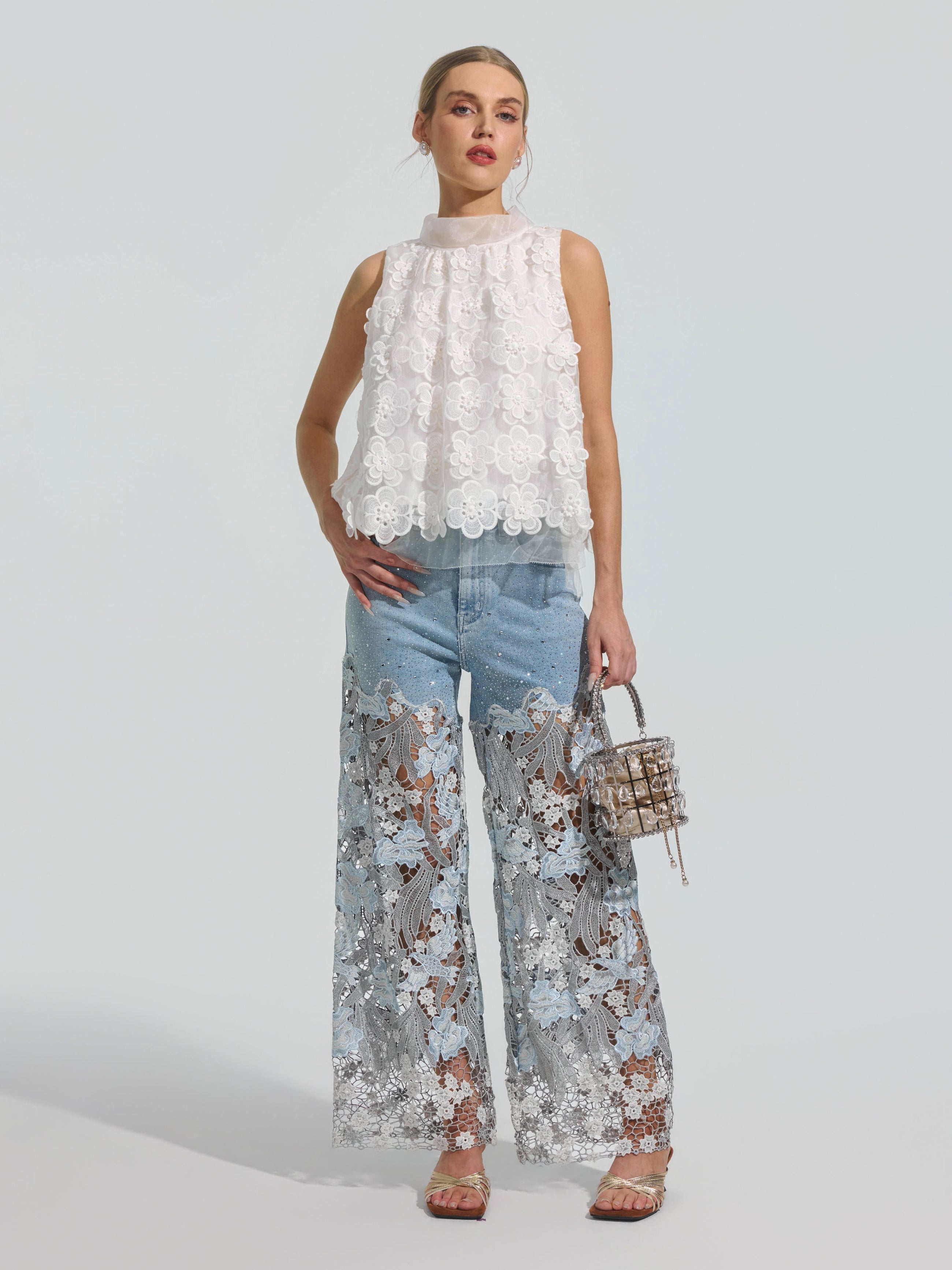 A woman stands against a plain background wearing Social Queen’s Floral Lace Patchwork Pants, styled with a sleeveless white top and a small handbag.