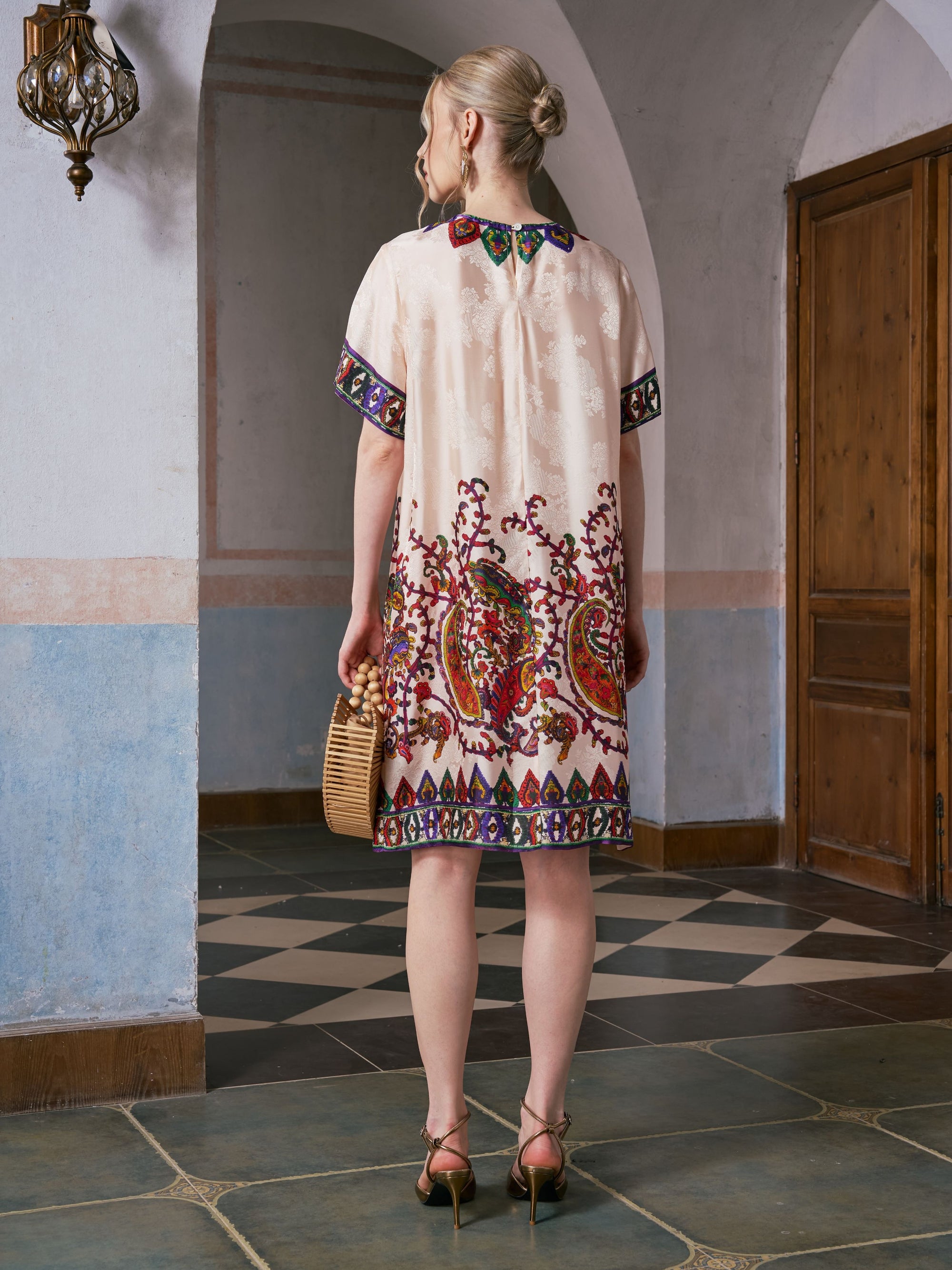 A woman stands indoors facing away, wearing the Social Queen Paisley Garden Print Jacquard Silk Dress, paired with a wicker handbag and heeled sandals.