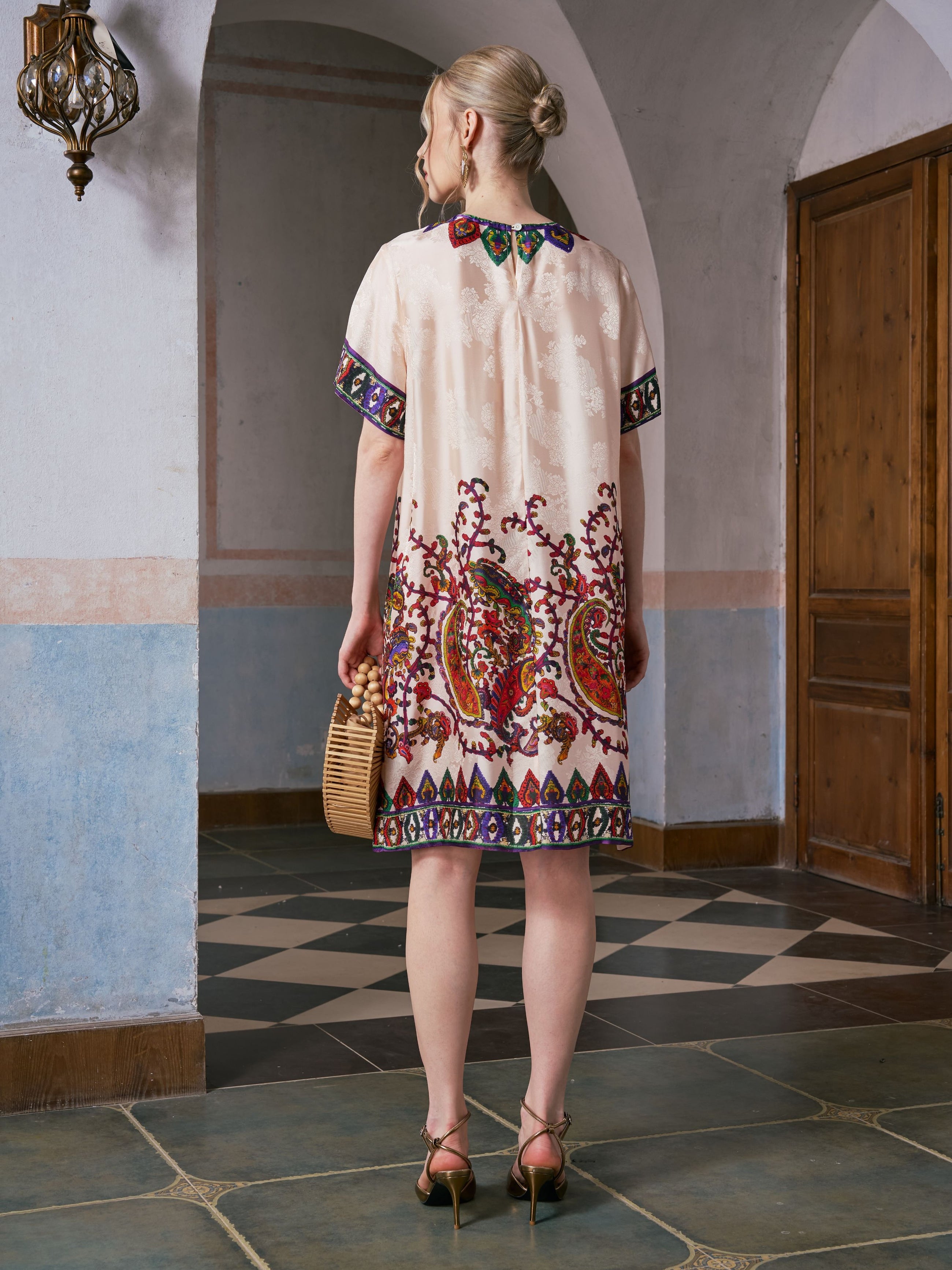 A woman stands indoors facing away, wearing the Social Queen Paisley Garden Print Jacquard Silk Dress, paired with a wicker handbag and heeled sandals.