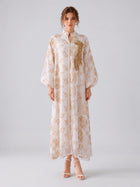 A woman stands against a plain background wearing the Social Queen Leaf Embroidered Beaded Dress—a long, white linen-like maxi dress featuring gold leaf embroidery and long sleeves—paired with white high-heeled sandals.