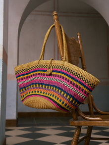 A Colorful Striped Woven Tote Bag by Social Queen, crafted from natural ramie fiber and adorned with vibrant patterns, hangs on the back of a wooden chair in a tiled, arched hallway.
