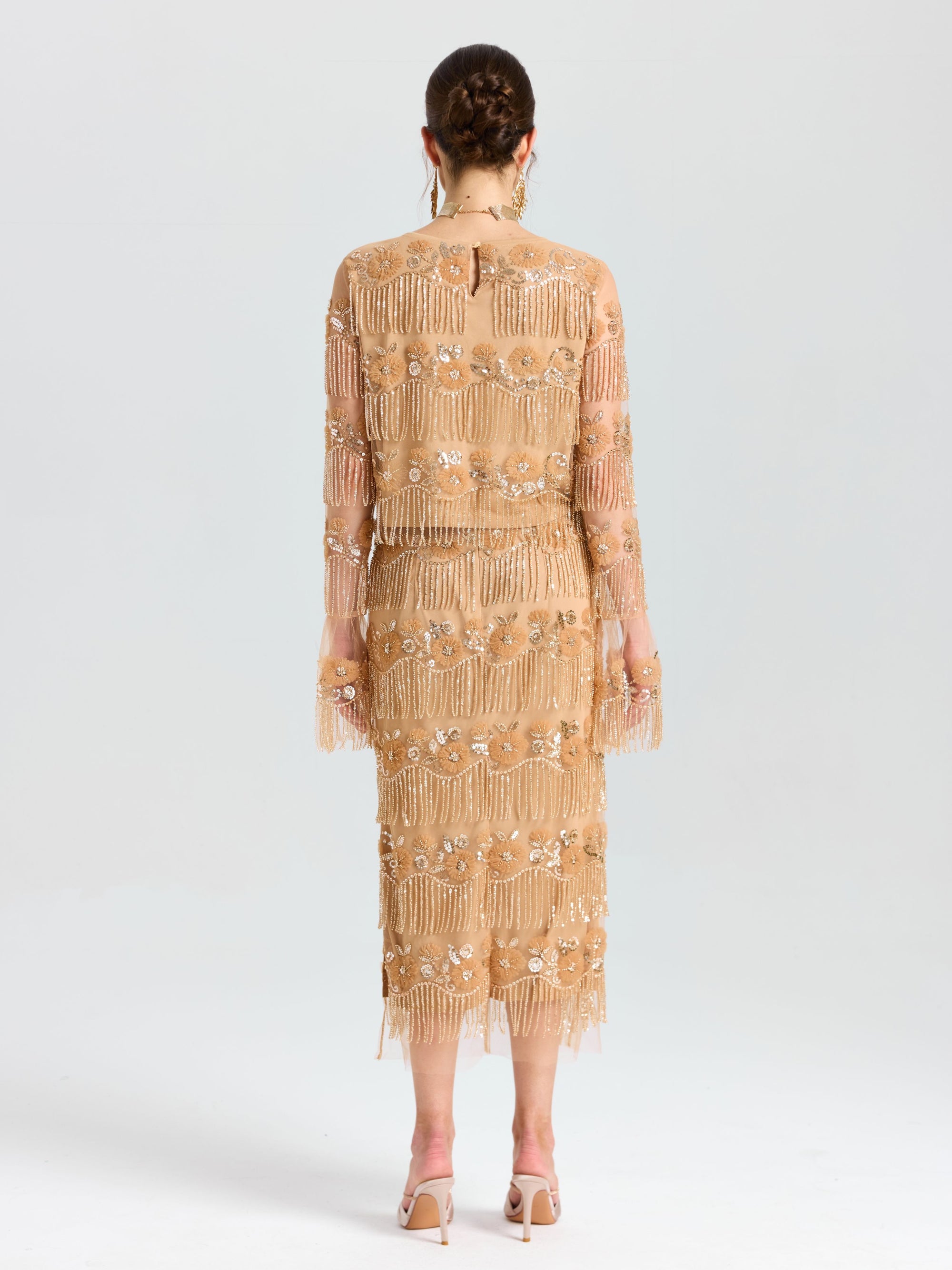 A woman faces away, wearing the Social Queen Beaded Fringe Slit Midi Skirt in a nude floral embroidery with beaded fringe. Her midi skirt features mesh accents, paired with heeled sandals and a braided updo.