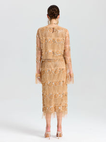 A woman faces away, wearing the Social Queen Beaded Fringe Slit Midi Skirt in a nude floral embroidery with beaded fringe. Her midi skirt features mesh accents, paired with heeled sandals and a braided updo.