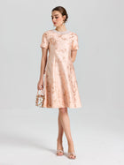 A woman wears the Social Queen Pearl Embellished Jacquard Dress, featuring a floral pattern and pearl-accented neckline. She pairs it with a small handbag and open-toe heels, standing against a plain background.