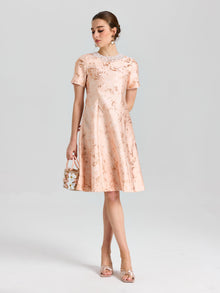 A woman wears the Social Queen Pearl Embellished Jacquard Dress, featuring a floral pattern and pearl-accented neckline. She pairs it with a small handbag and open-toe heels, standing against a plain background.