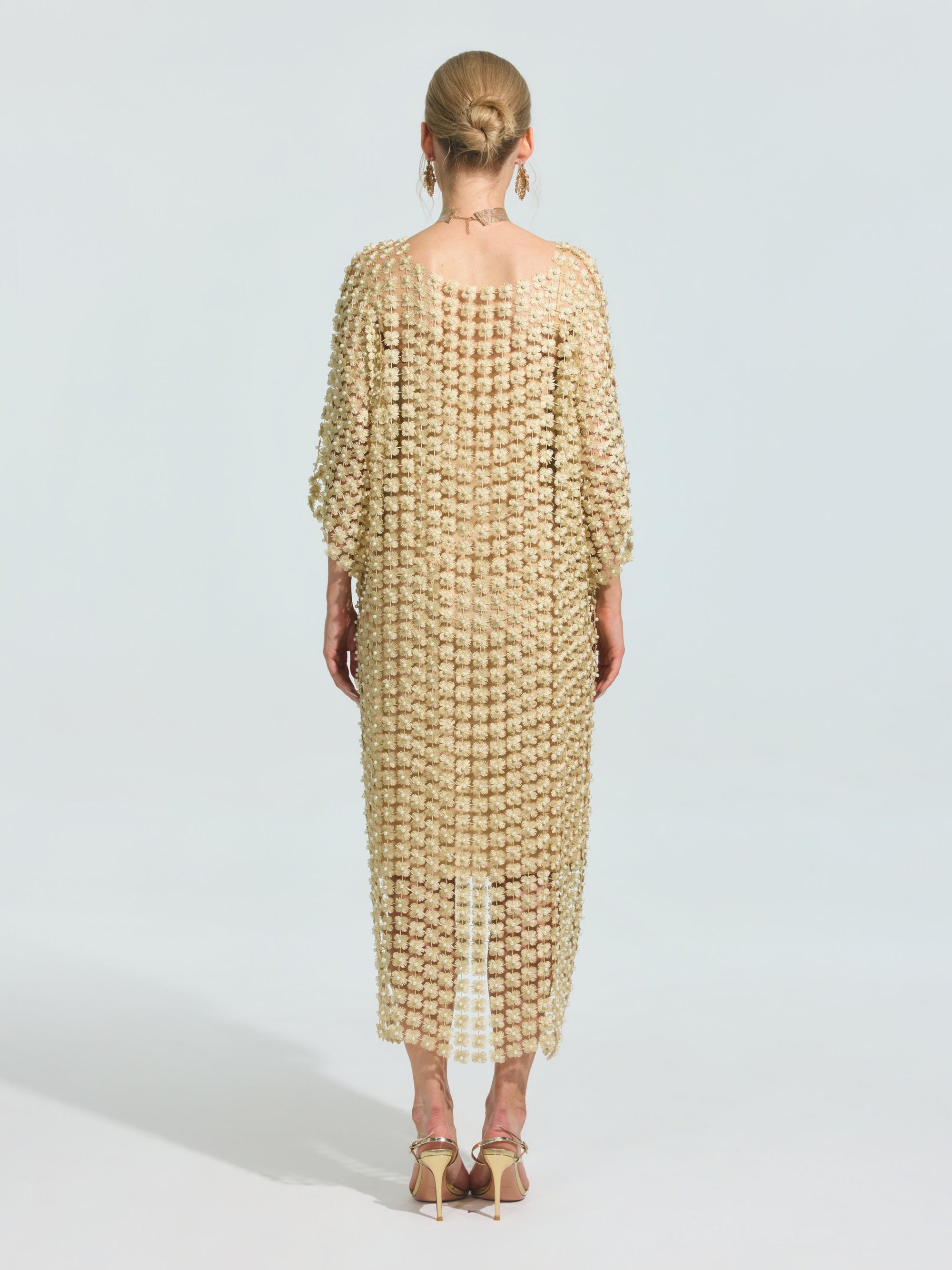 A woman stands facing away, wearing the Pearl Embellished Floral Overlay Dress (Slip Dress Included) by Social Queen, distinguished by its loose fit and elegant detailing. Her hair is in a bun, and she pairs the look with heeled sandals.