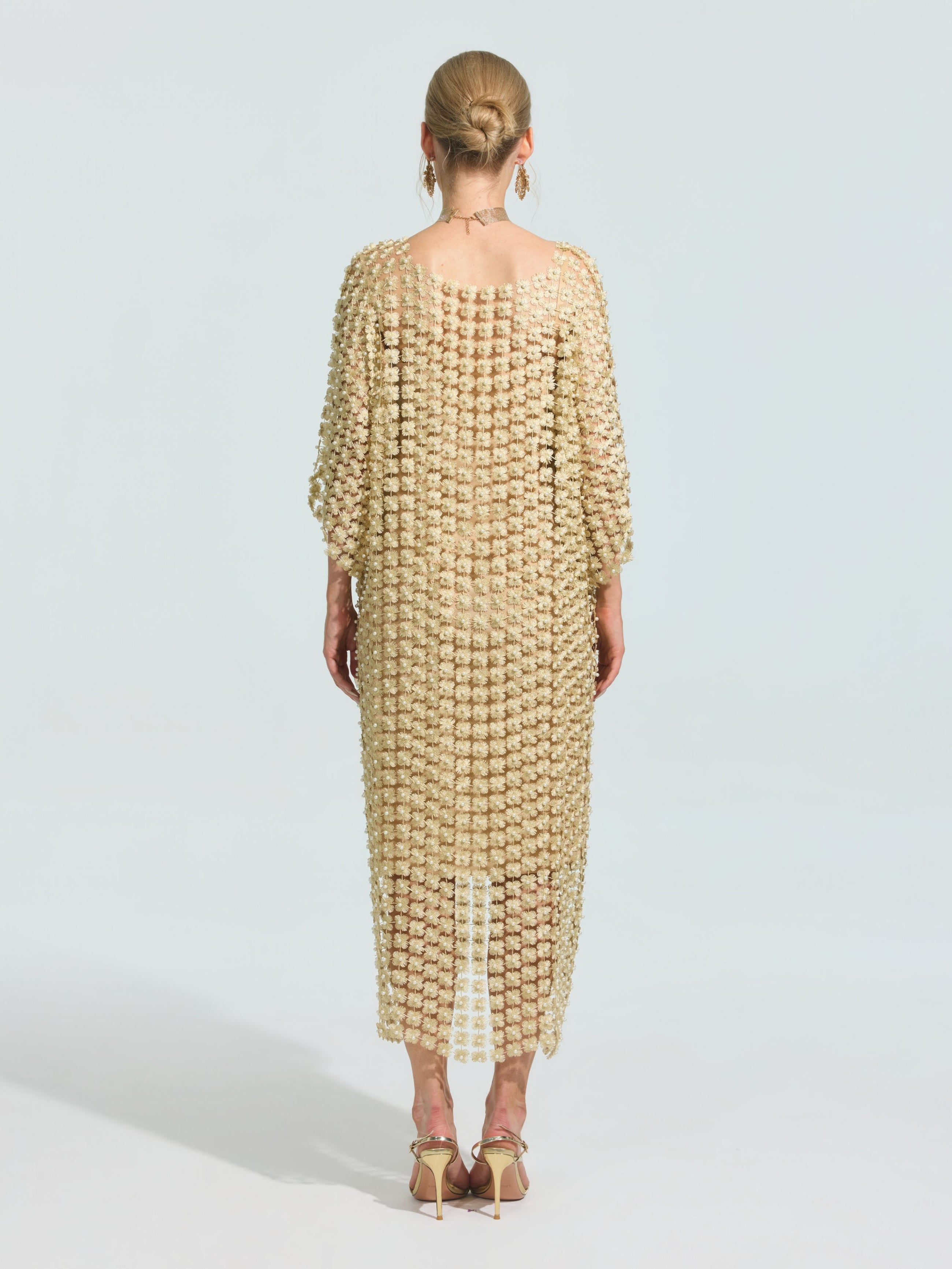 A woman stands facing away, wearing the Pearl Embellished Floral Overlay Dress (Slip Dress Included) by Social Queen, distinguished by its loose fit and elegant detailing. Her hair is in a bun, and she pairs the look with heeled sandals.