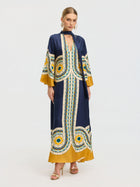 A woman models the Social Queen Moroccan Print Silk Caftan Dress in blue, yellow, and white with gold heels and hoop earrings, standing against a plain white background.