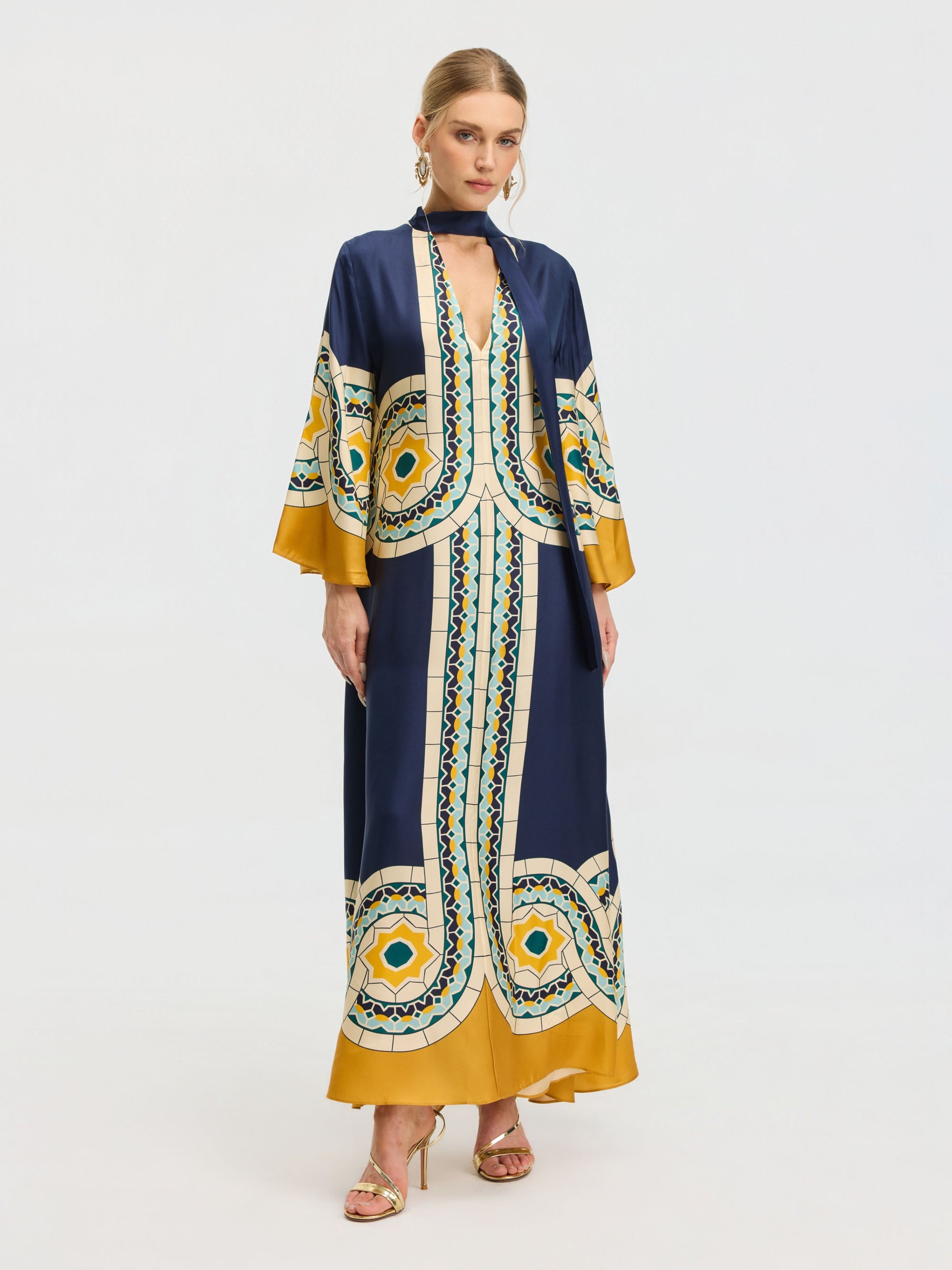 A woman models the Social Queen Moroccan Print Silk Caftan Dress in blue, yellow, and white with gold heels and hoop earrings, standing against a plain white background.