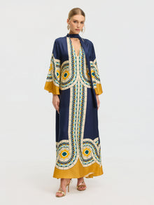 A woman models the Social Queen Moroccan Print Silk Caftan Dress in blue, yellow, and white with gold heels and hoop earrings, standing against a plain white background.