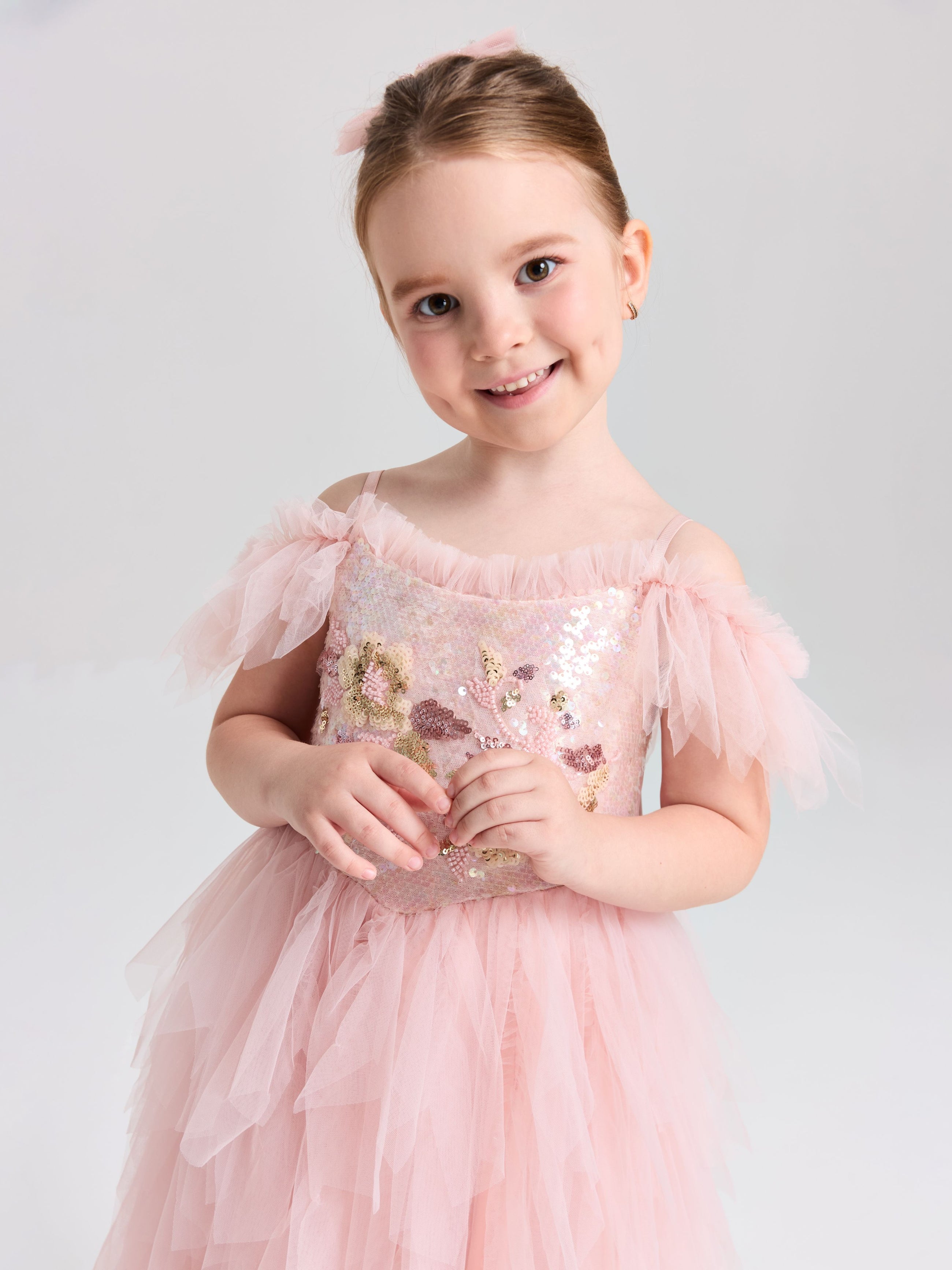 A young girl smiles against a plain, light background wearing the Social Queen Girls Floral Sequin Tulle Dress in light pink with ruffled details.