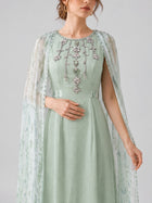 A woman wearing the Social Queen Beaded Printed Cape Chiffon Dress, adorned with floral embellishments and shimmering beaded details, featuring a sheer patterned cape, stands against a plain light background.
