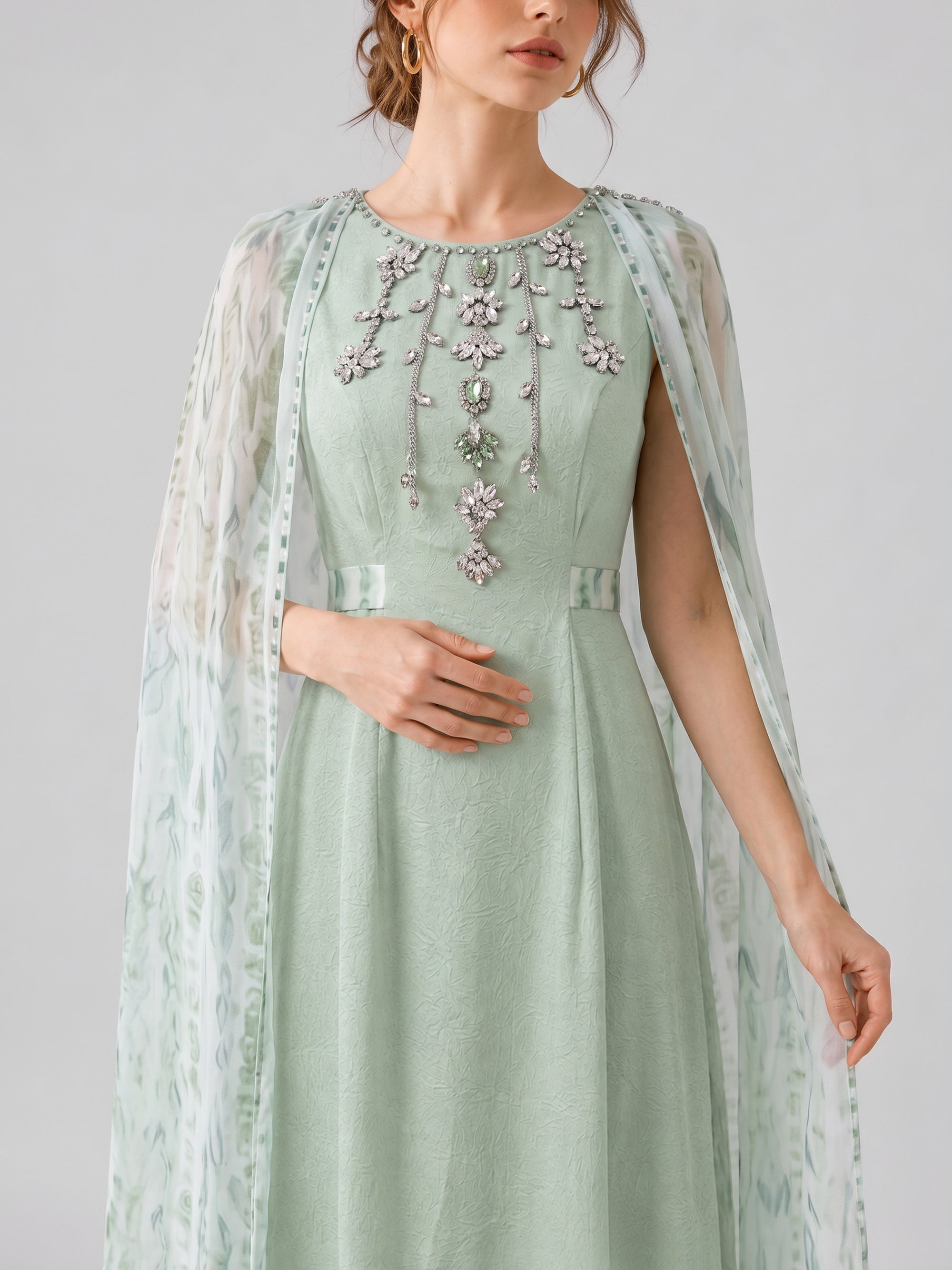 A woman wearing the Social Queen Beaded Printed Cape Chiffon Dress, adorned with floral embellishments and shimmering beaded details, featuring a sheer patterned cape, stands against a plain light background.