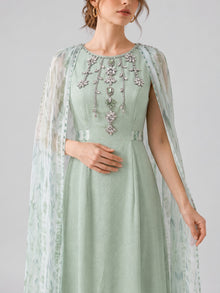 A woman wearing the Social Queen Beaded Printed Cape Chiffon Dress, adorned with floral embellishments and shimmering beaded details, featuring a sheer patterned cape, stands against a plain light background.
