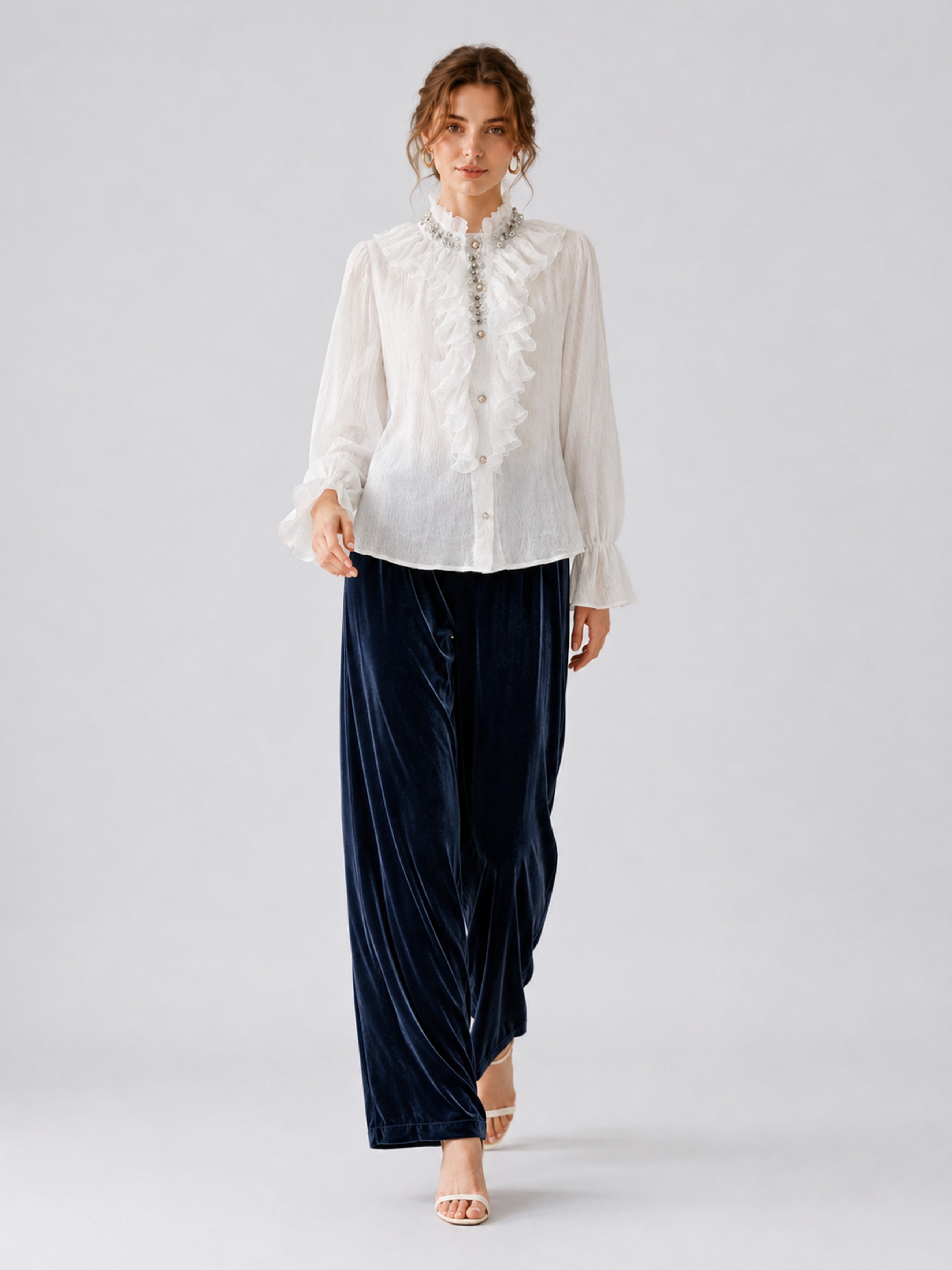 A woman stands against a plain light background wearing the Social Queen Silk Velvet Wide-Leg Pants in navy blue, styled with a white ruffled blouse and heeled sandals.