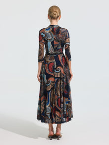 A woman stands with her back to the camera wearing the Social Queen Paisley Print Knit Dress (belt included), a long-sleeved, ankle-length stretch knit dress with a colorful paisley pattern, paired with low-heeled shoes.