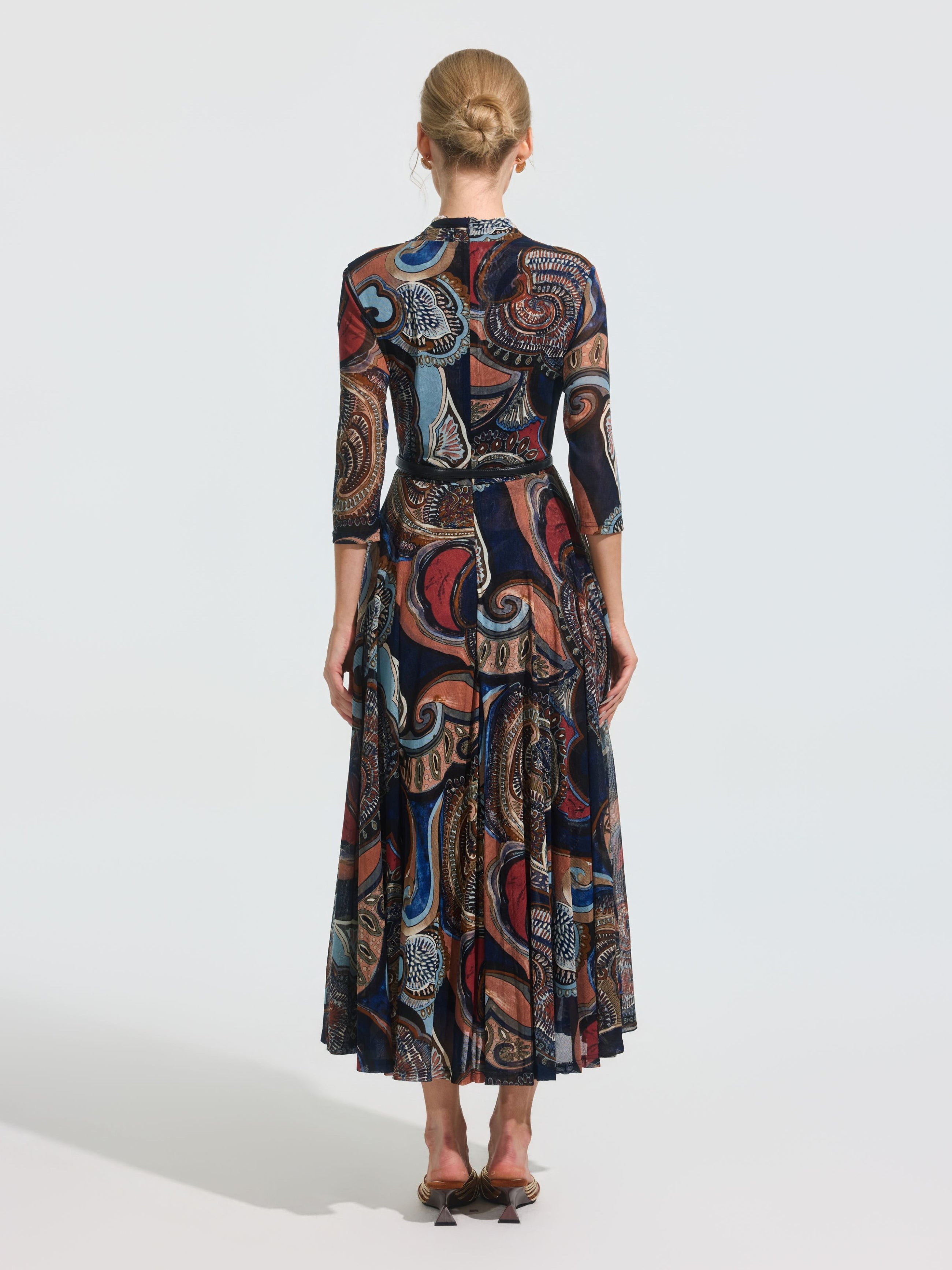 A woman stands with her back to the camera wearing the Social Queen Paisley Print Knit Dress (belt included), a long-sleeved, ankle-length stretch knit dress with a colorful paisley pattern, paired with low-heeled shoes.