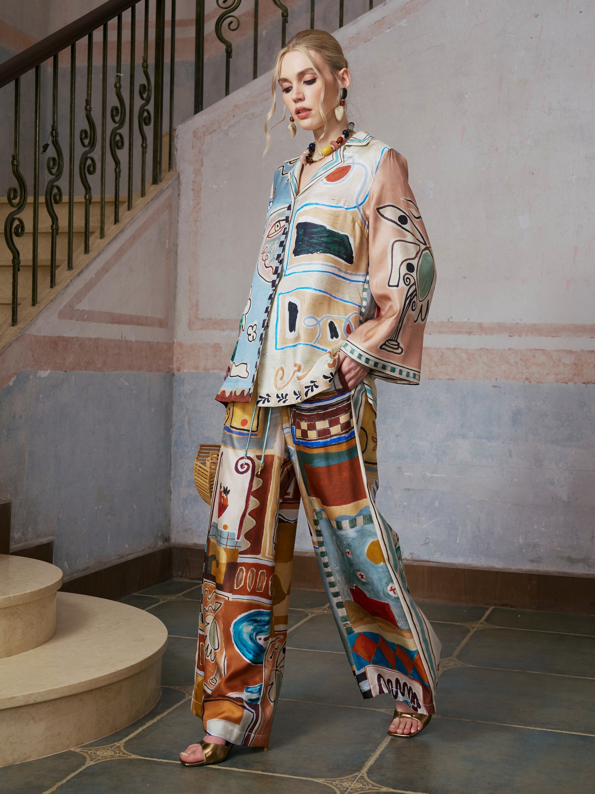 A woman stands on stone steps wearing the Abstract Print Mulberry Silk Shirt by Social Queen, a loose-fitting resort wear piece featuring colorful abstract patterns, and holds a woven bag. A stair railing and textured wall appear in the background.