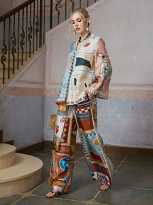A woman stands on stone steps wearing the Abstract Print Mulberry Silk Shirt by Social Queen, a loose-fitting resort wear piece featuring colorful abstract patterns, and holds a woven bag. A stair railing and textured wall appear in the background.