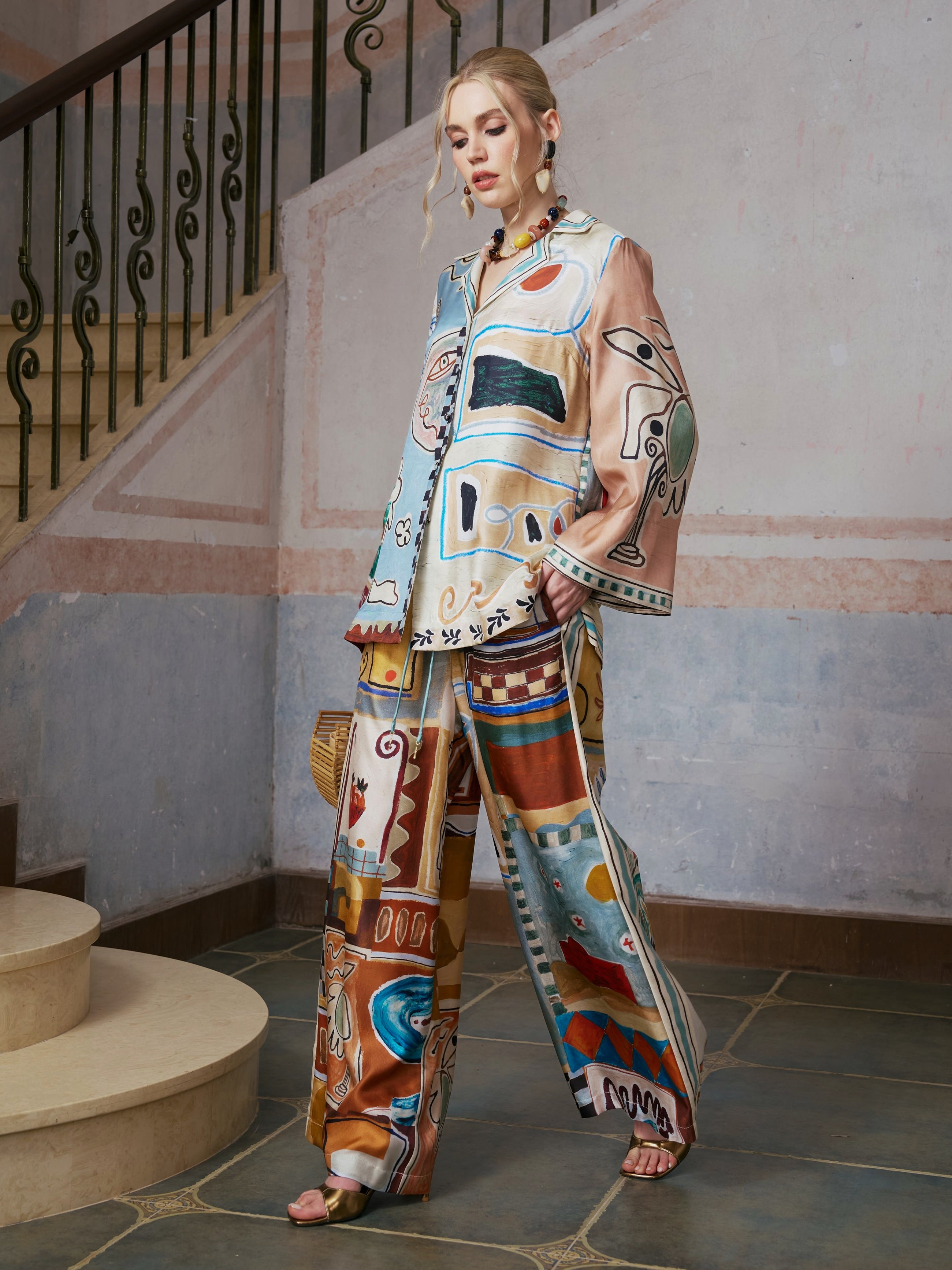 A woman stands on stone steps wearing the Abstract Print Mulberry Silk Shirt by Social Queen, a loose-fitting resort wear piece featuring colorful abstract patterns, and holds a woven bag. A stair railing and textured wall appear in the background.