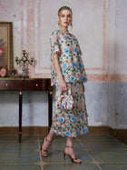 A woman wearing the Social Queen 3D Floral Cutwork Lace Top, adorned with delicate blue 3D floral appliqués, holds a small patterned handbag while standing in a vintage room featuring a console table and decorative accents.