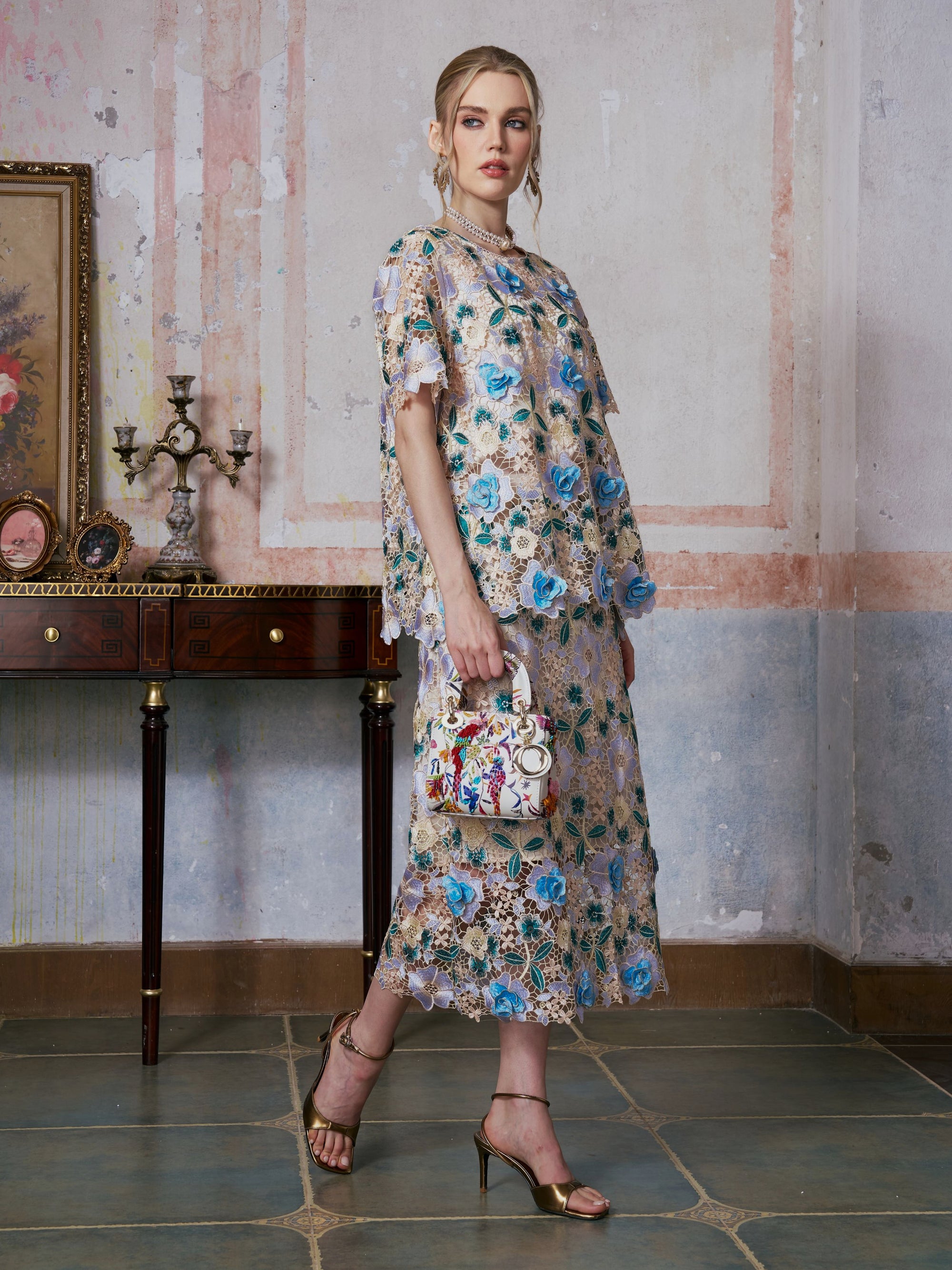 A woman wearing the Social Queen 3D Floral Cutwork Lace Top, adorned with delicate blue 3D floral appliqués, holds a small patterned handbag while standing in a vintage room featuring a console table and decorative accents.