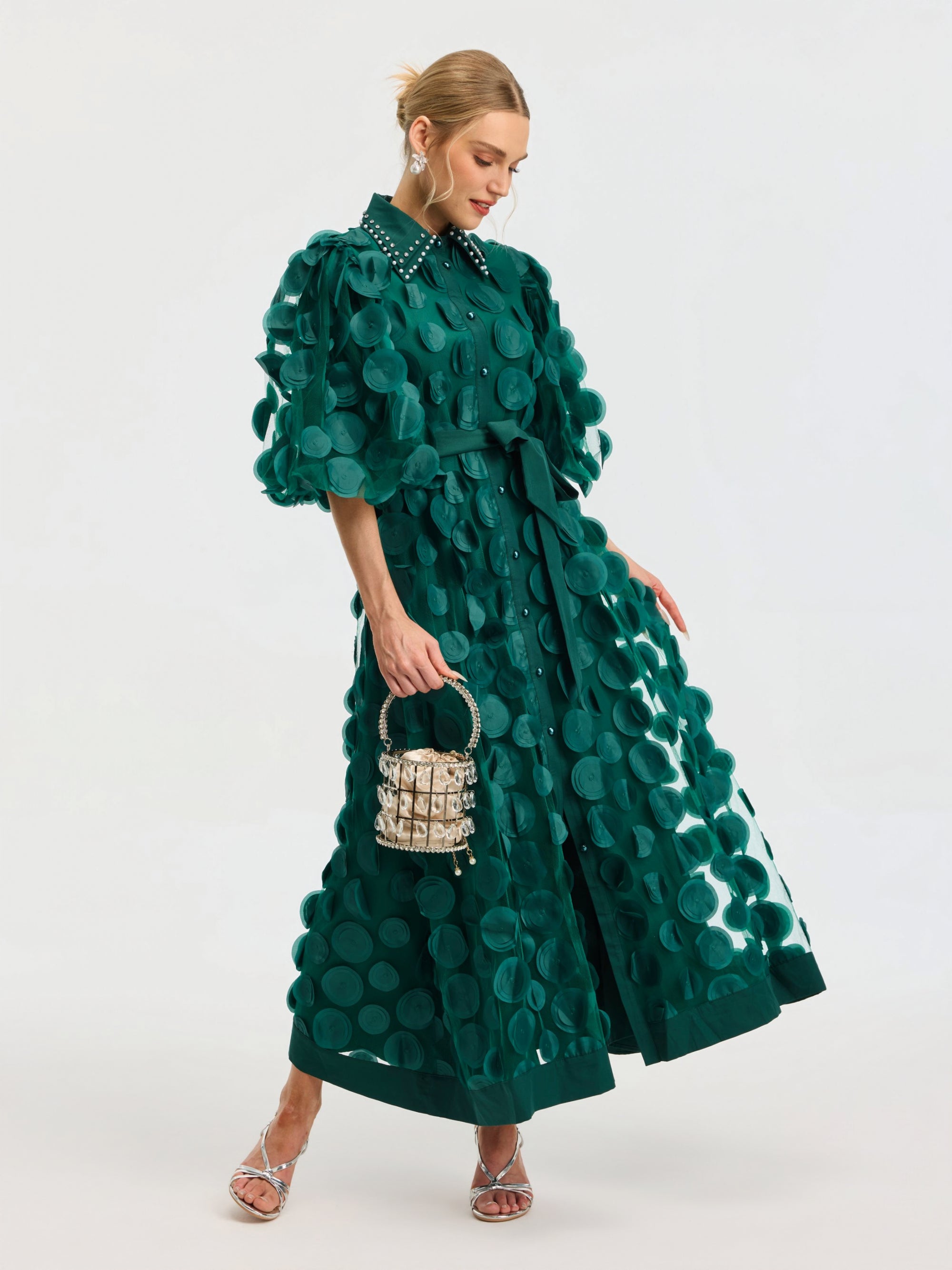A woman wears the Social Queen Pearl Embellished Appliqué Dress, featuring green textured fabric with large polka dots and crystal pearl details. She pairs it with a small purse and silver strappy heels against a plain background.