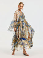 A woman models the Social Queen Byzantine Print Silk Caftan Dress, adorned with blue, gold, and tan patterns and a crystal-embellished neckline, styled with strappy sandals against a plain background.