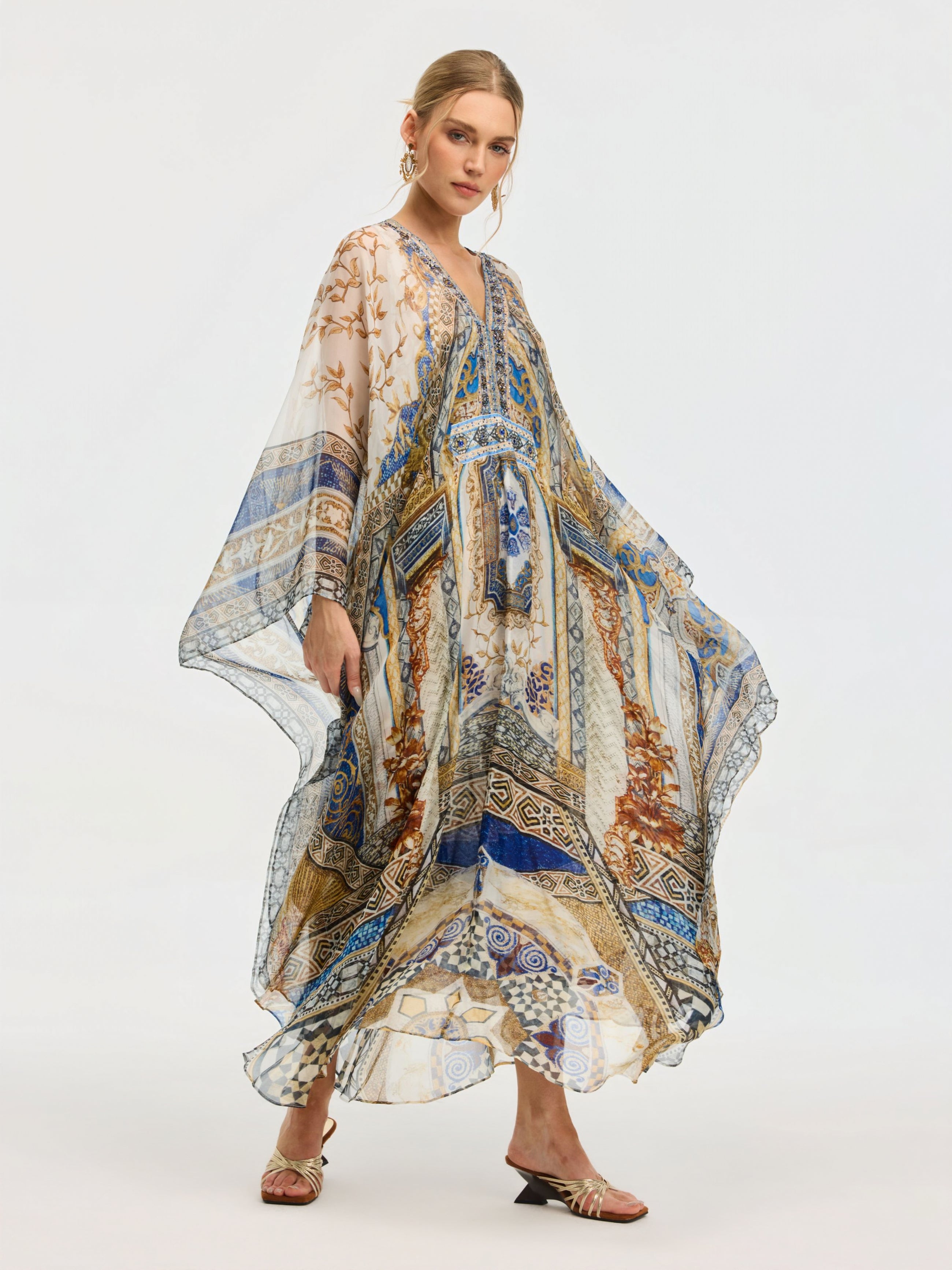 A woman models the Social Queen Byzantine Print Silk Caftan Dress, adorned with blue, gold, and tan patterns and a crystal-embellished neckline, styled with strappy sandals against a plain background.