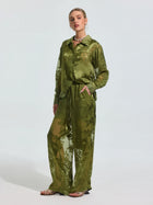 A woman stands with one hand in her pocket, wearing Social Queen's Botanical Burnout Satin Wide-Leg Pants and a matching green floral shirt, styled with open-toe heels and a necklace.