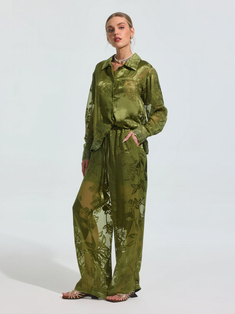 A woman stands with one hand in her pocket, wearing Social Queen's Botanical Burnout Satin Wide-Leg Pants and a matching green floral shirt, styled with open-toe heels and a necklace.