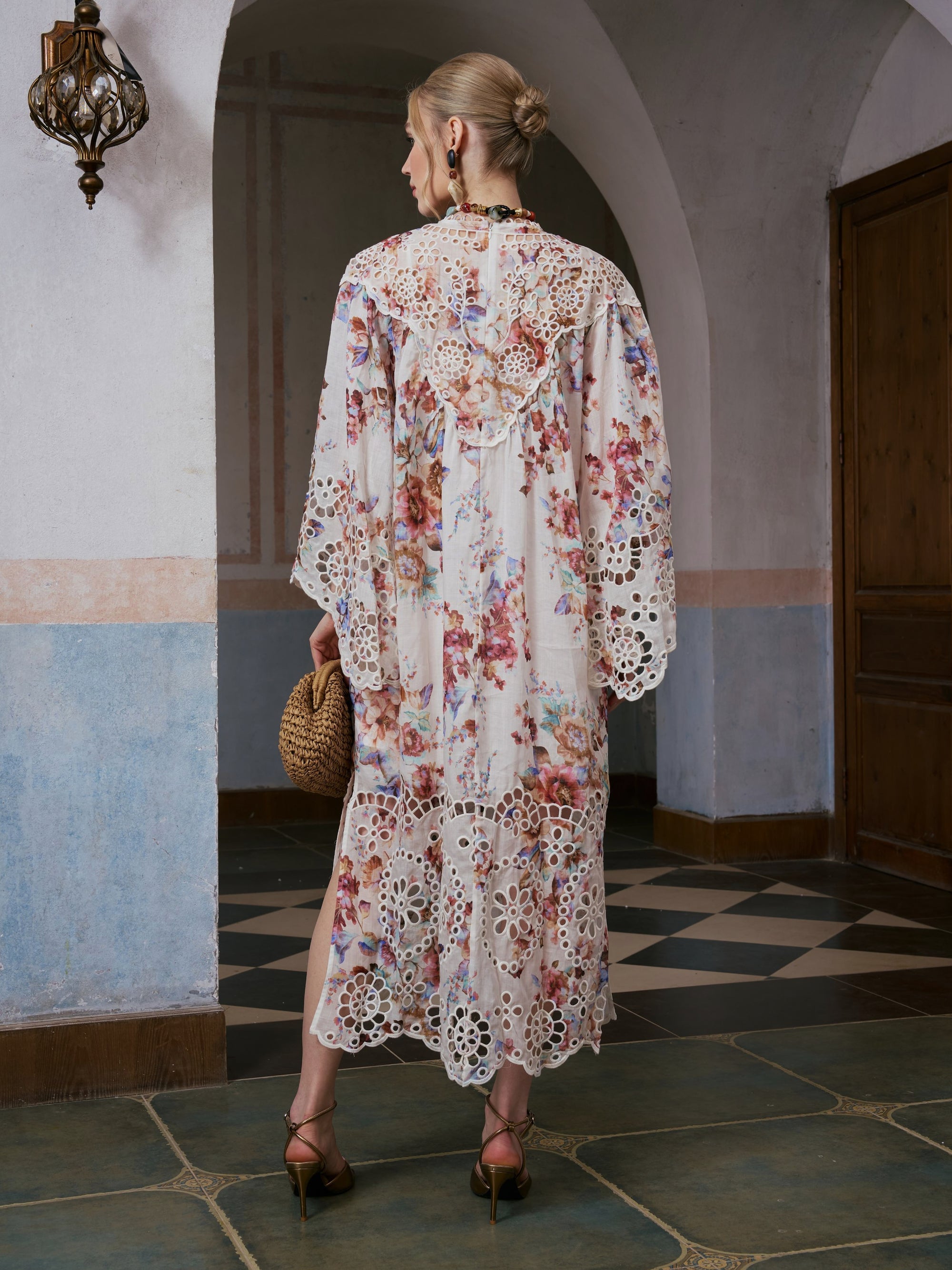 A woman stands indoors on a tiled floor, facing away from the camera, wearing the Social Queen Floral Print Cutwork Linen Dress and heels, and carrying a woven handbag.