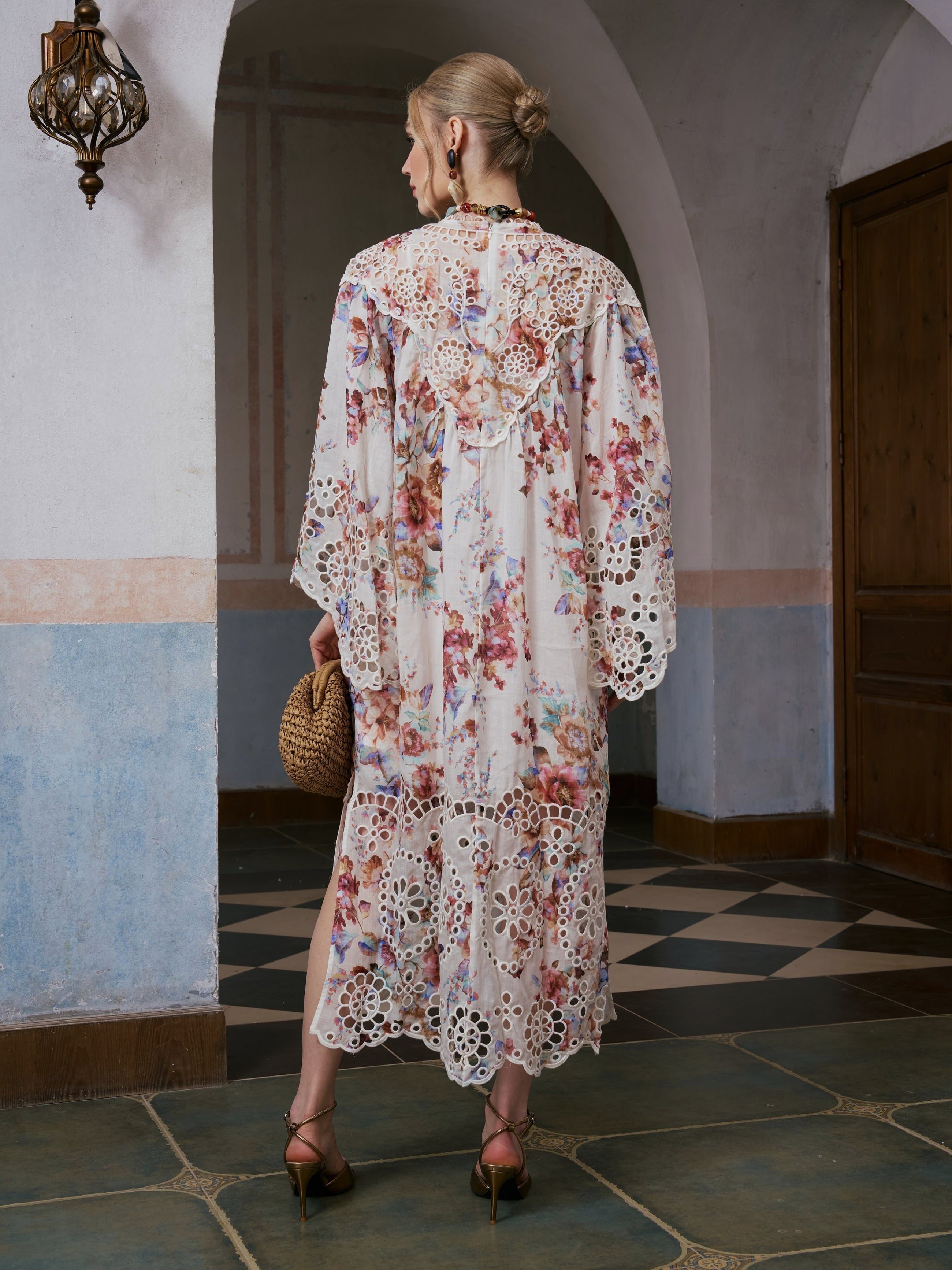 A woman stands indoors on a tiled floor, facing away from the camera, wearing the Social Queen Floral Print Cutwork Linen Dress and heels, and carrying a woven handbag.