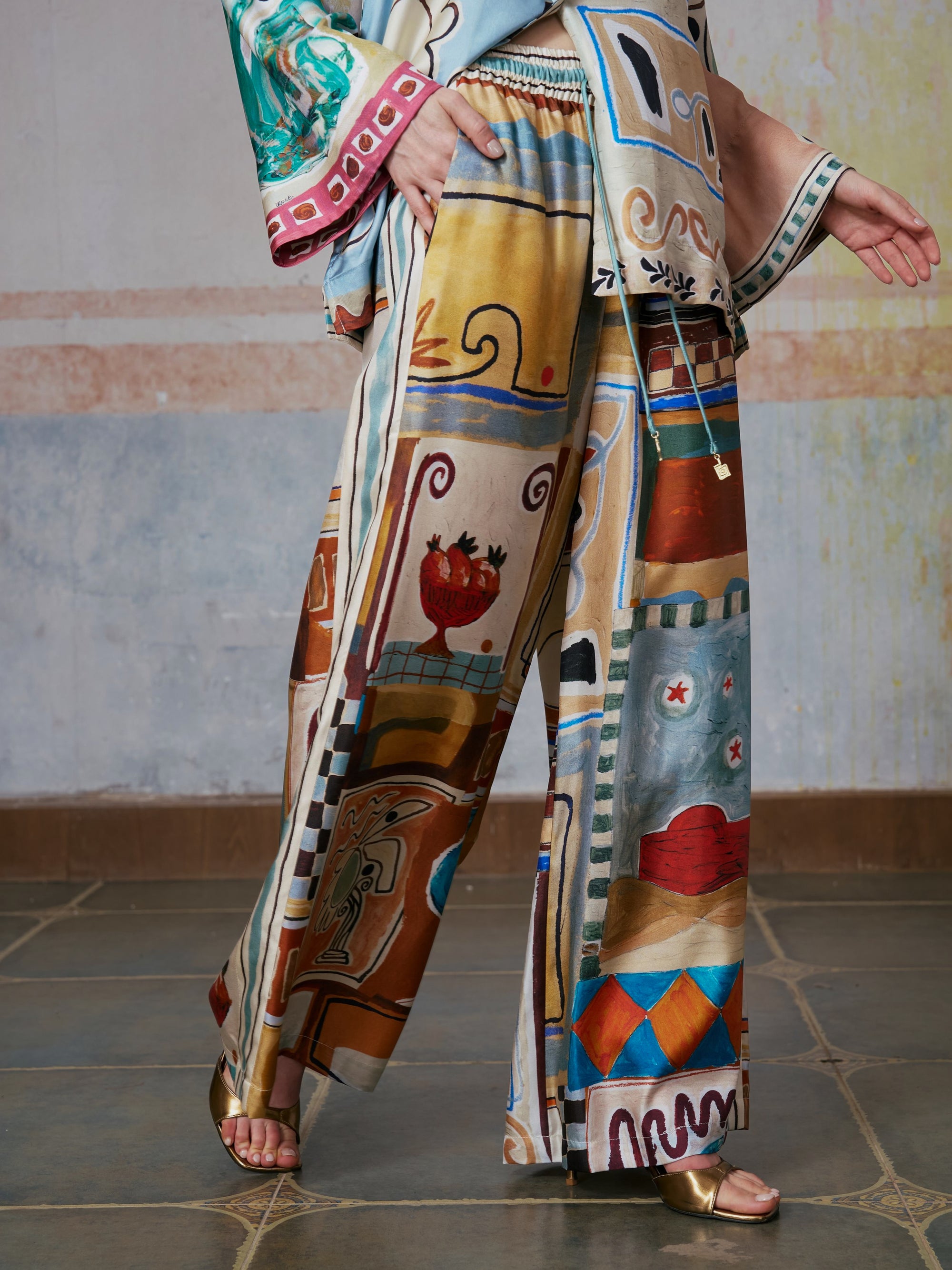 Wearing Social Queen’s Abstract Print Mulberry Silk Pants and a matching loose top adorned with vibrant, abstract patterns, the person stands indoors on a tiled floor, one foot forward and arms gently lifted.