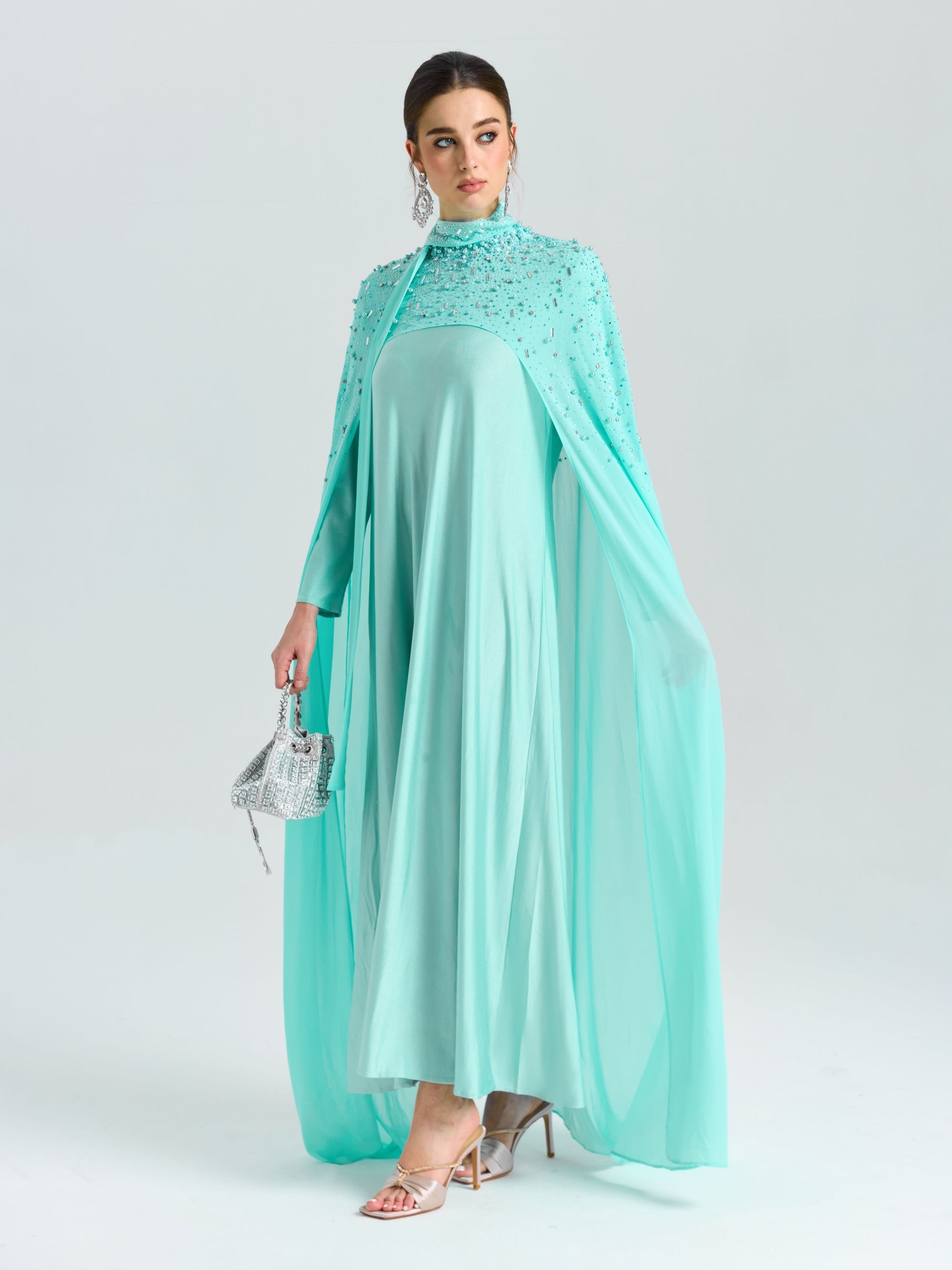 A woman wears the Social Queen Beaded Cape Scarf Gown, featuring flowing light blue chiffon and delicate embellishments, holding a small silver handbag against a plain light background.