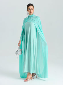 A woman wears the Social Queen Beaded Cape Scarf Gown, featuring flowing light blue chiffon and delicate embellishments, holding a small silver handbag against a plain light background.