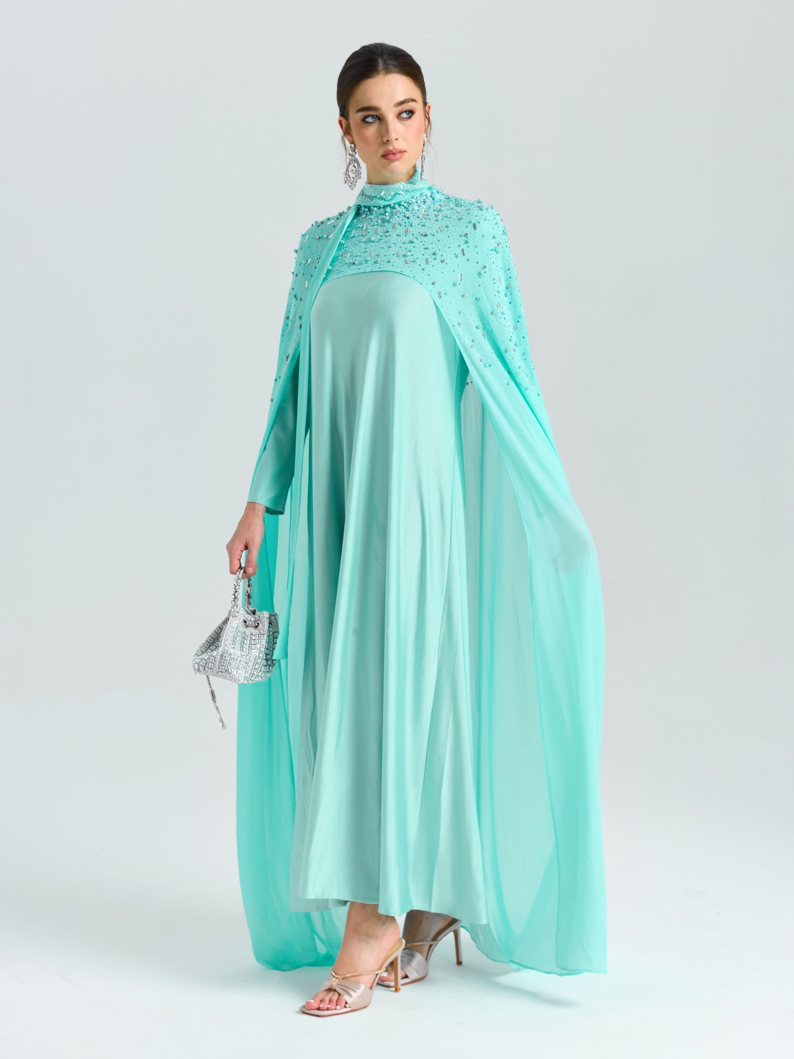 A woman wears the Social Queen Beaded Cape Scarf Gown, featuring flowing light blue chiffon and delicate embellishments, holding a small silver handbag against a plain light background.